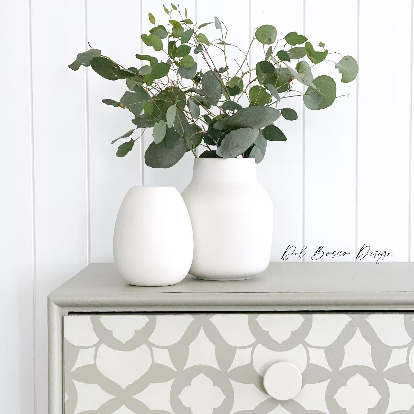 close up of Two white vases with greenery on a patterned dresser against a white paneled wall featuring white wooden drawer knobs.