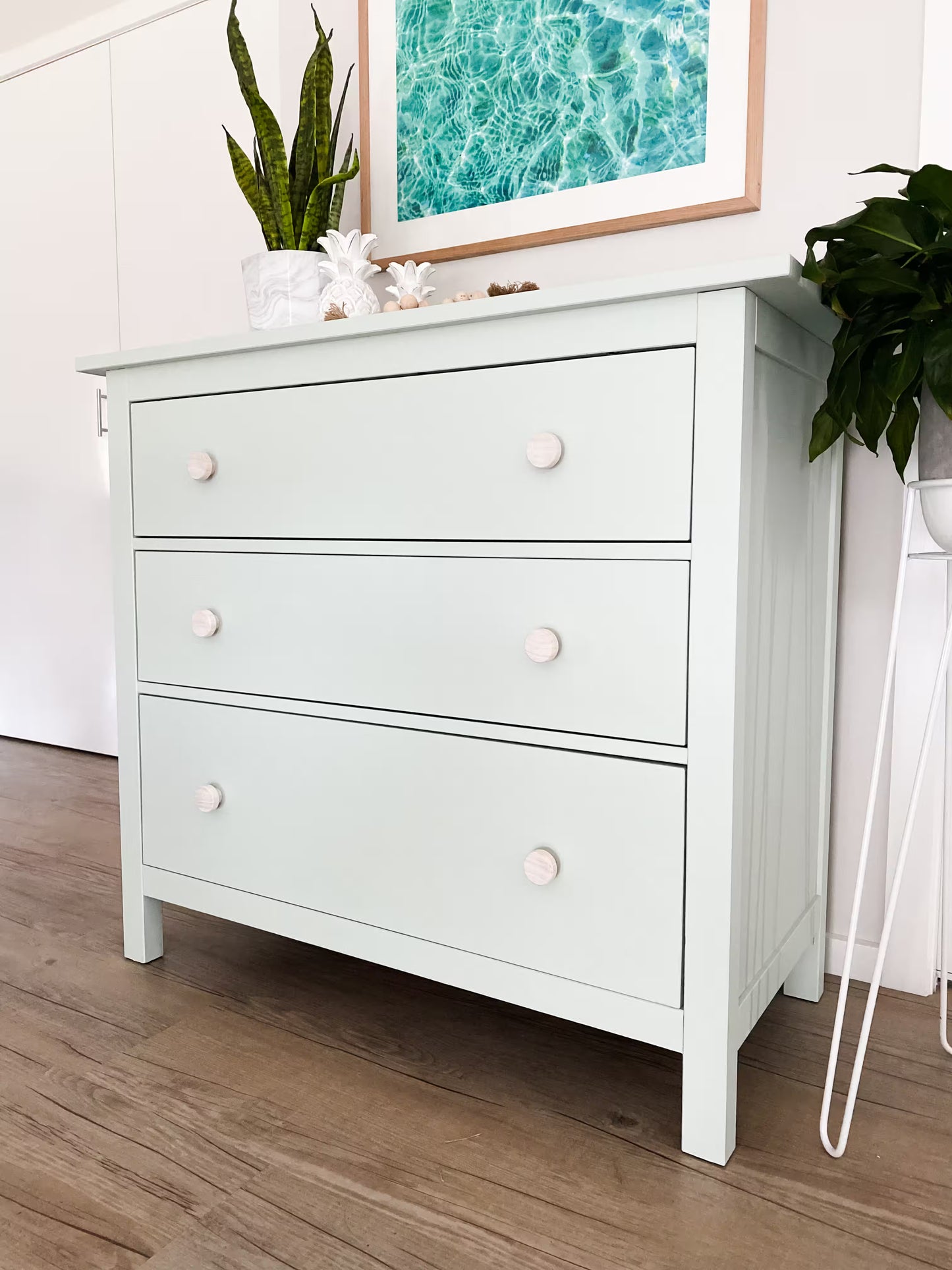 White dresser with white wooden knobs in a coastal styled room with plants and artwork on the wall.