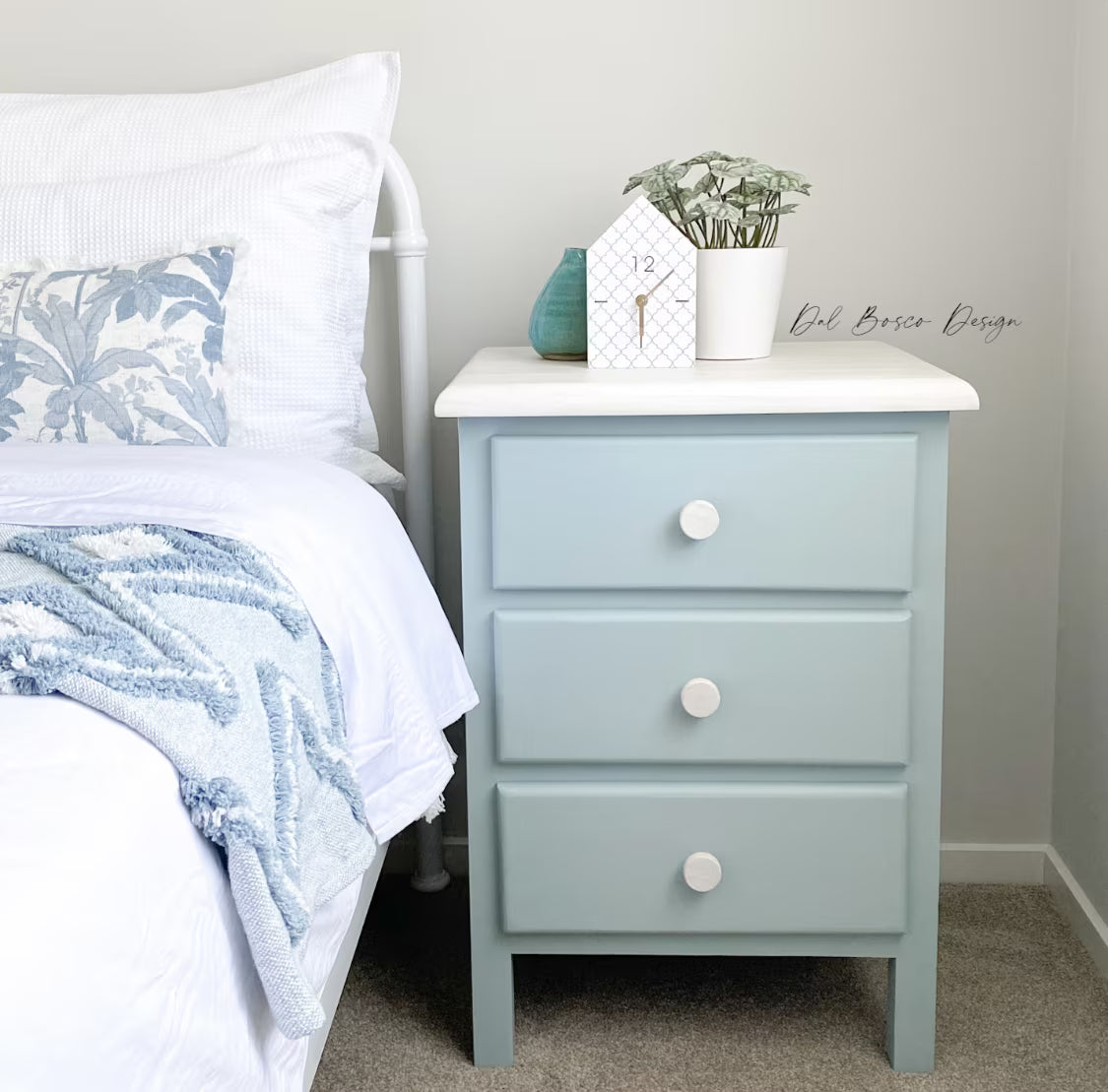 Light blue nightstand with white drawer knobs next to a bed with white bedding and a decorative pillow.