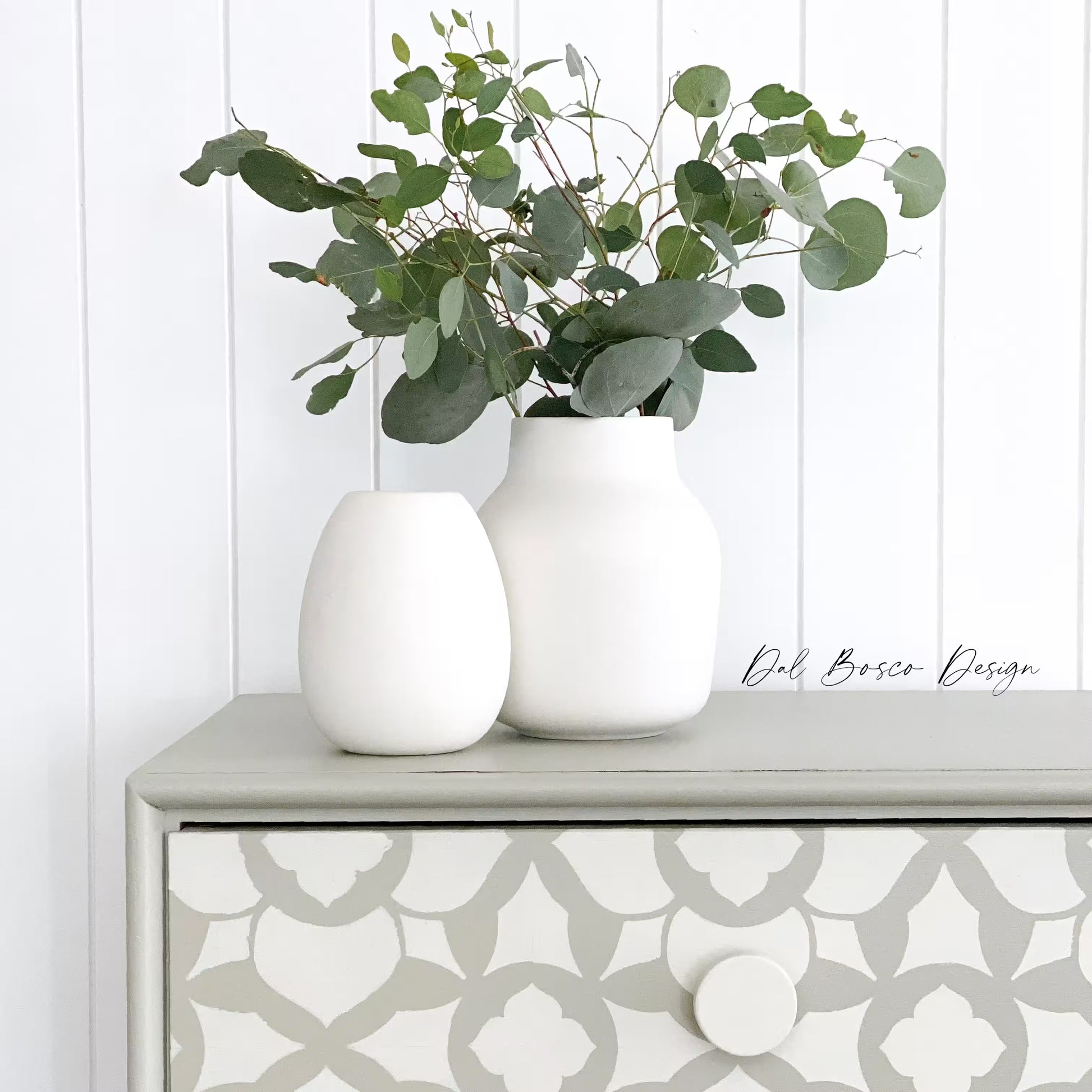 close up of Two white vases with greenery on a patterned dresser against a white paneled wall featuring white wooden drawer knobs.