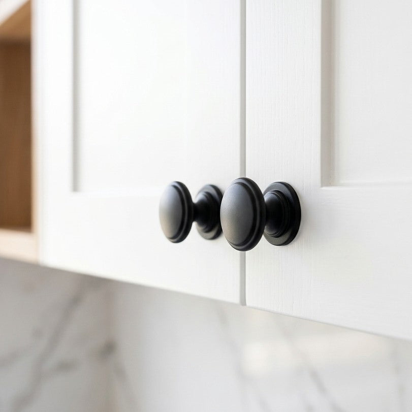 Close-up architectural photograph of a pair of classic matte black round cabinet knobs installed on white timber wood-grain Shaker style overhead kitchen cupboard doors.