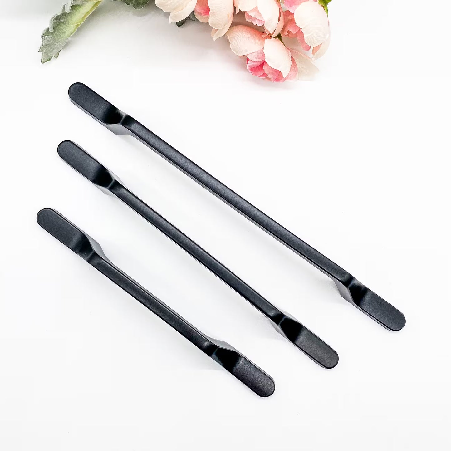 Three black drawer handles on a white surface with pink flowers in the background