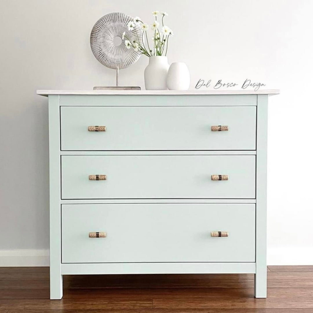 Pale green chest of drawers featuring rattan drawer handles against white wall on timber floor